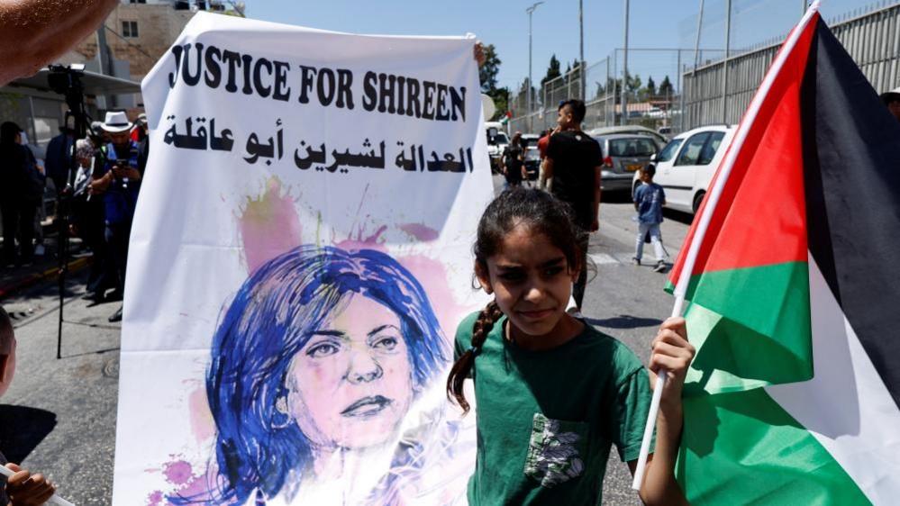 A young Palestinian girl holds up a Palestinian flag and a poster that says Justice for Shireen with a drawing of the Palestinian-American journalist.