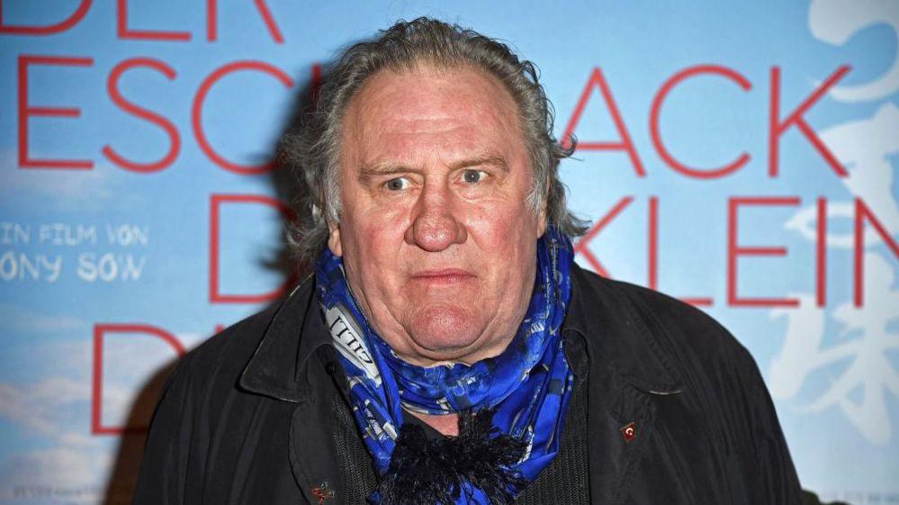 Gerard Depardieu appears at a film premiere wearing a blue scarf in front of a film poster