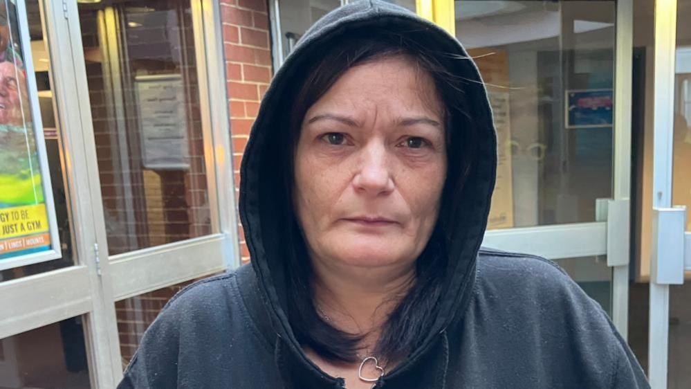 A woman with dark hair and dark hoodie, with the hood up. She is stood outside the leisure centre.