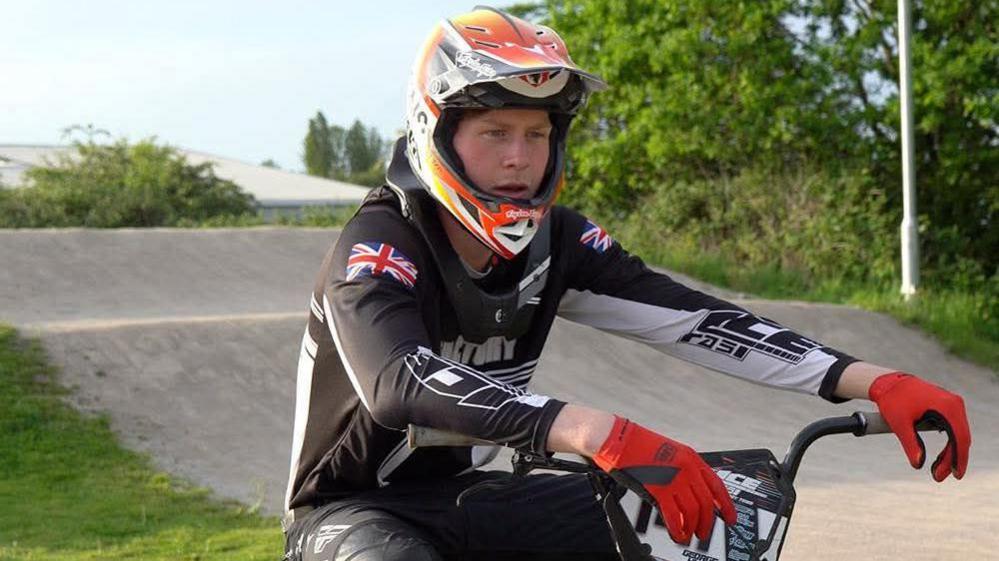George Hunt sits on his BMX bike on a track. He is wearing cycling gear and red gloves with a helmet. There are trees around the track.