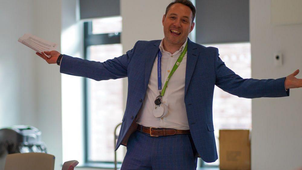 Ed looking happy in a blue suit and white shirt with his arms outstretched wearing a lanyard