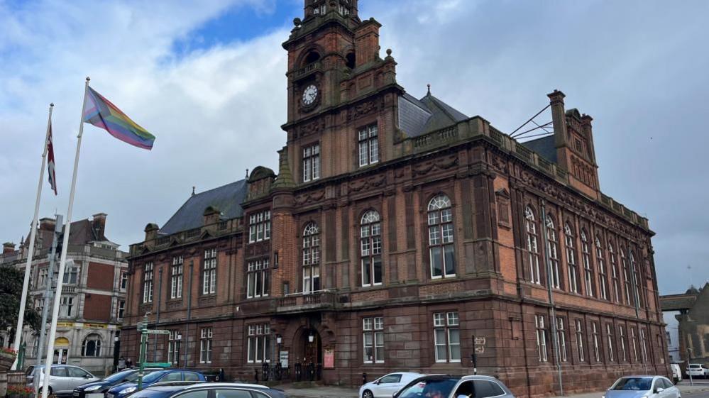 Great Yarmouth Town Hall, an imposing neo-classical building with large sash windows. A Progress Pride flag is flying outside the front of the building, alongside a Union Jack flag. 