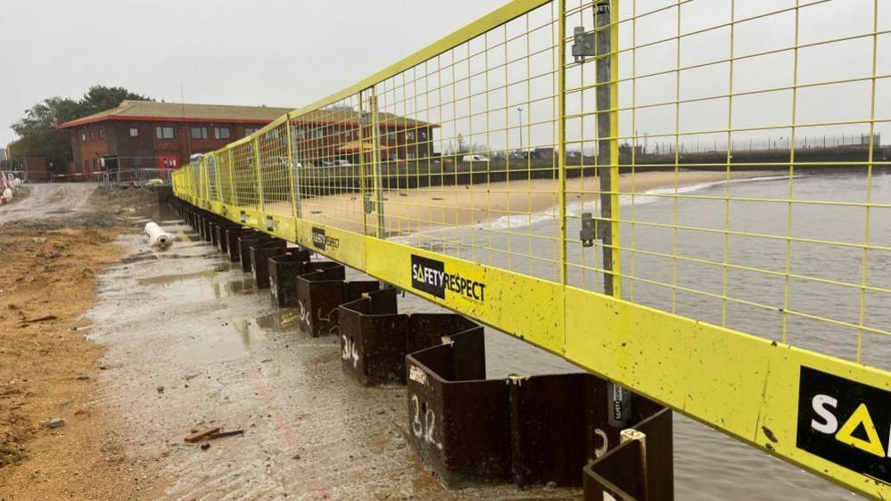 Sheet steel piling and concrete with yellow fencing and a beach on the river in the distance