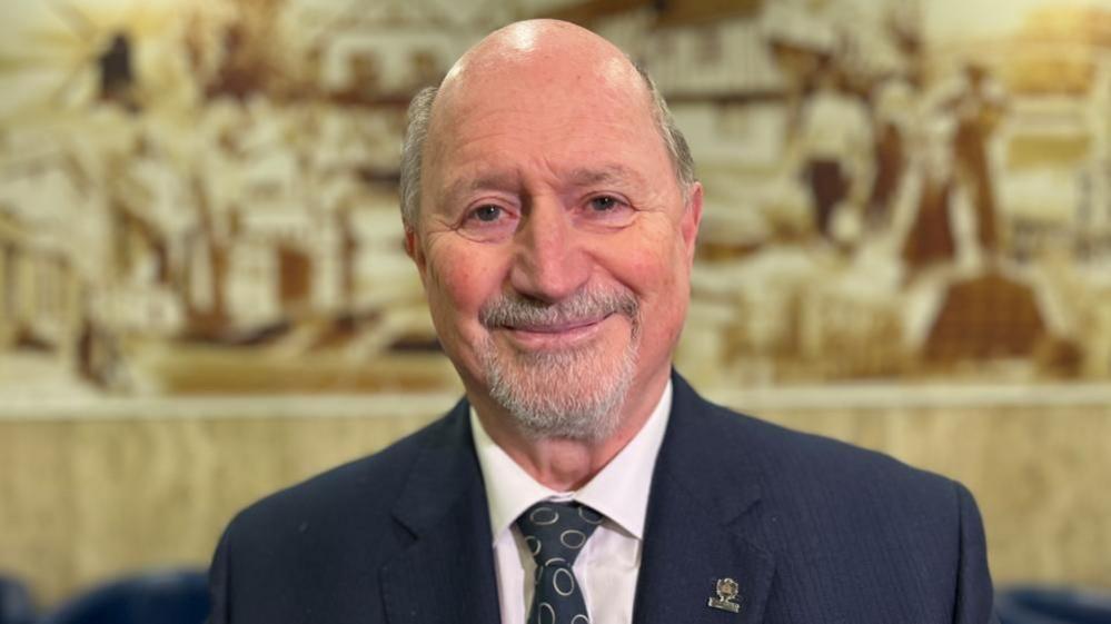 Jim Weir with short grey hair and geard, wearing a dark grey jacket, light-coloured shirt and black tie with a yellow circle pattern. He is standing in front of decorative stone edging.