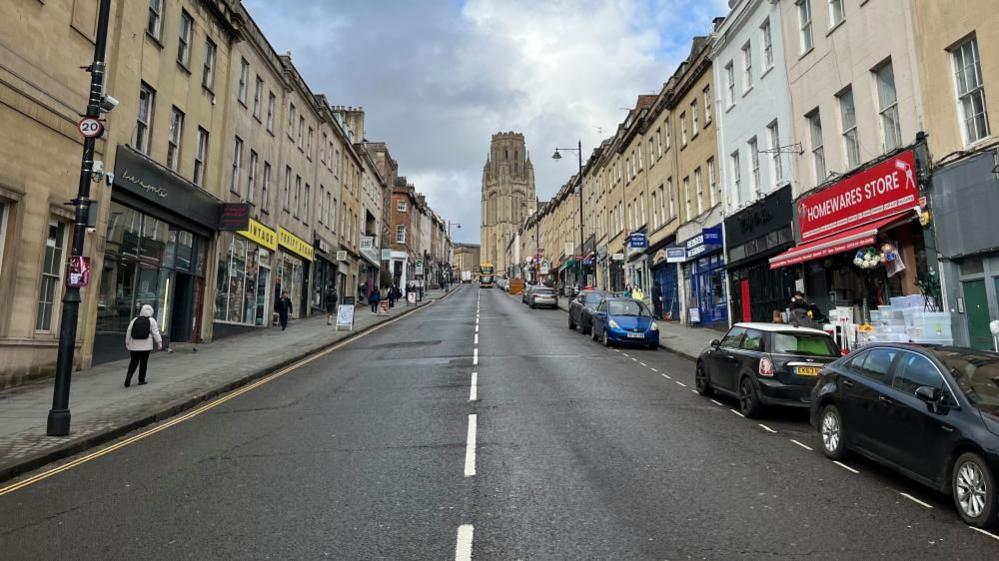 An empty road looking up a hill. It is lined by shops and bars and there are cars parked on one side. A large tower - Wills Memorial Building - can be seen at the top of the route.