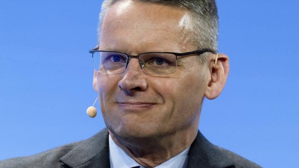 A close-up of John Pettigrew, who wears a suit and glasses and a small beige microphone that is hooked from his left ear, around the back of his head to stick out subtly to the right of his mouth