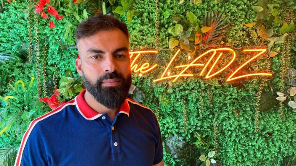 A bearded man in a blue polo shirt with red trim stands in front of a floral display.