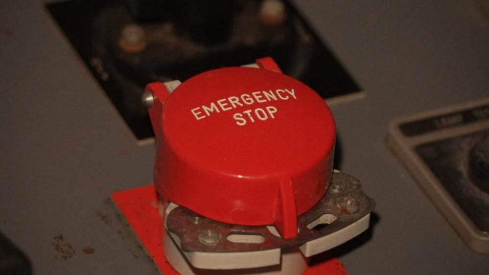 A large red emergency stop button, with the words engraved in white on the top. The button is set within a grey control panel.