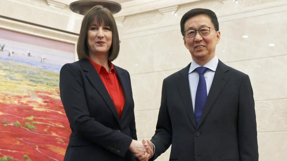 Reeves, wearing a black suit with red shirt, shakes the hand of Han Zheng, who wears a black suit with white shirt and blue tie