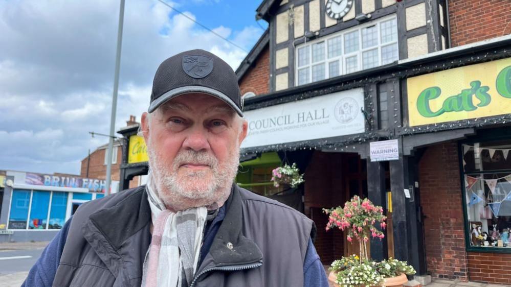 Kevin Wood, chairman of Caister Parish Council, wearing a blue cap, grey jacket and multi-coloured scarf. He is standing outside the council hall, which is a red brick building.