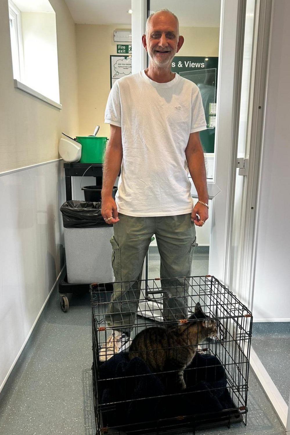 Ian Haskins, wearing a white T-shirt and green grey trousers, is standing with Ethan, who is in a crate, at his feet. Mr Haskins has white hair and a white beard and is smiling broadly.