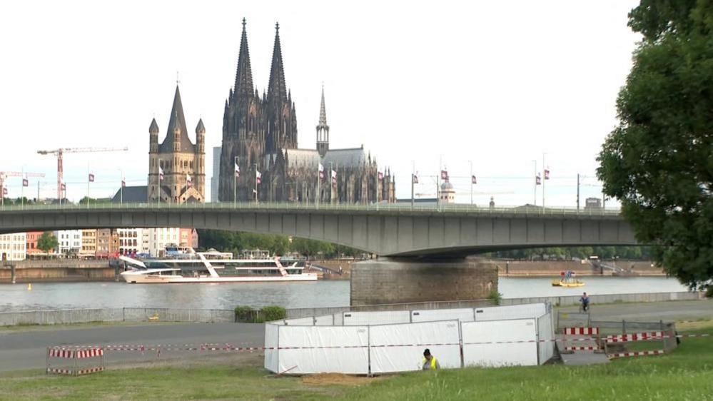 A person works by a cordoned off in a grassy area near a river, with Cologne Cathedral in the background