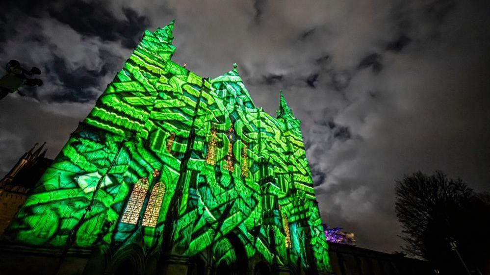 The facade of Salisbury Cathedral lit up for the Sarum Lights. Against a black and grey sky, a green maze pattern is projected onto the cathedral wall.