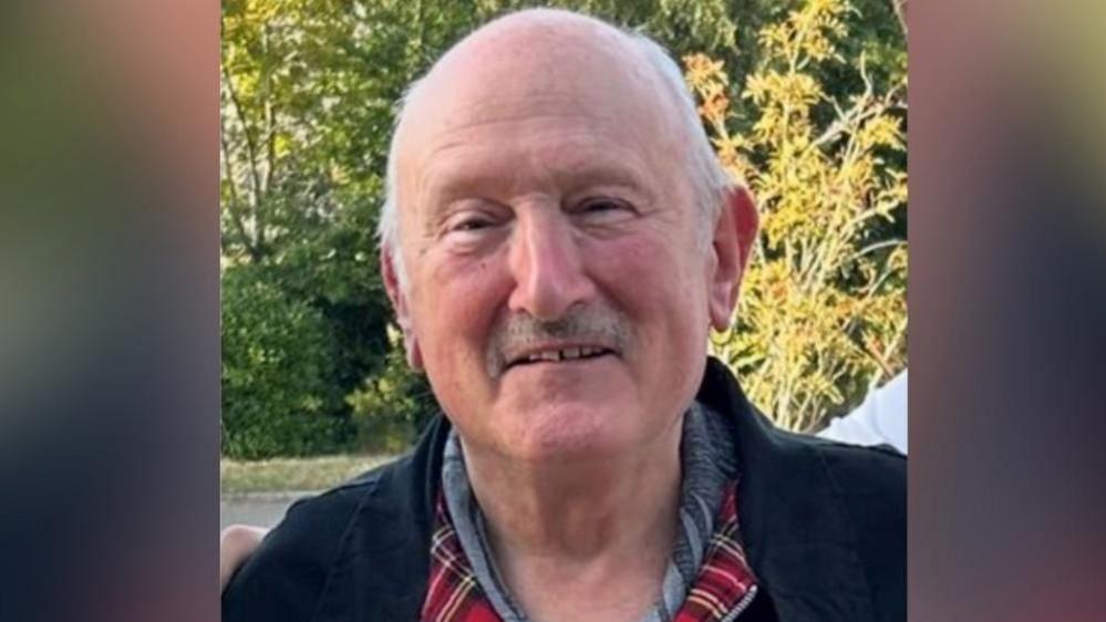 Terry McFadyen has greying short hair and a short moustache. He is wearing a black jacket with a red tartan lining and a shirt under. He is smiling. Trees are seen in the image behind where he is standing.