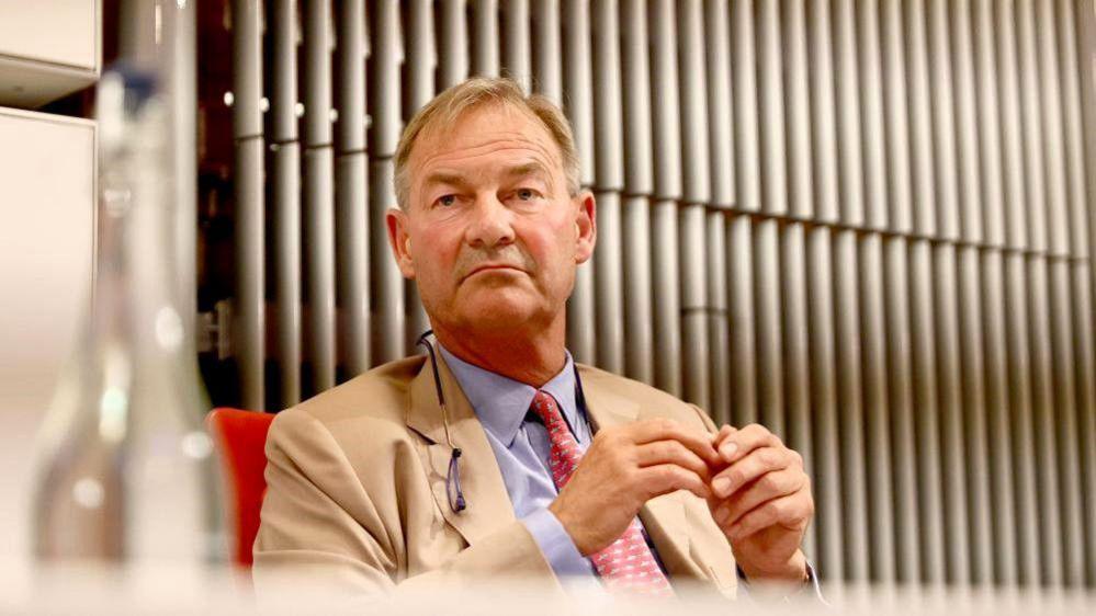 Stock photo of Rupert Lowe sat on a chair wearing a beige blazer, blue shirt and red tie. He is unsmiling and looking to the left of the camera.   