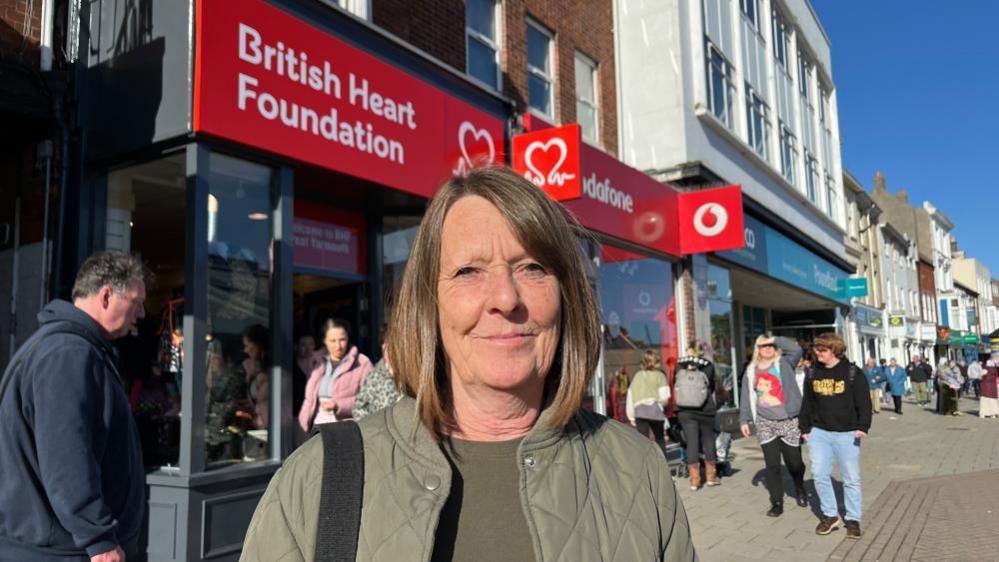 Jane Hare is photographed standing outside the British Heart Foundation shop, which had a red facia sign with white lettering. She is wearing an olive green quilted jacket and a matching knitted top. She has a black bag strap over her right shoulder and she has shoulder length light brown hair. Other people are walking in the vicinity around her.