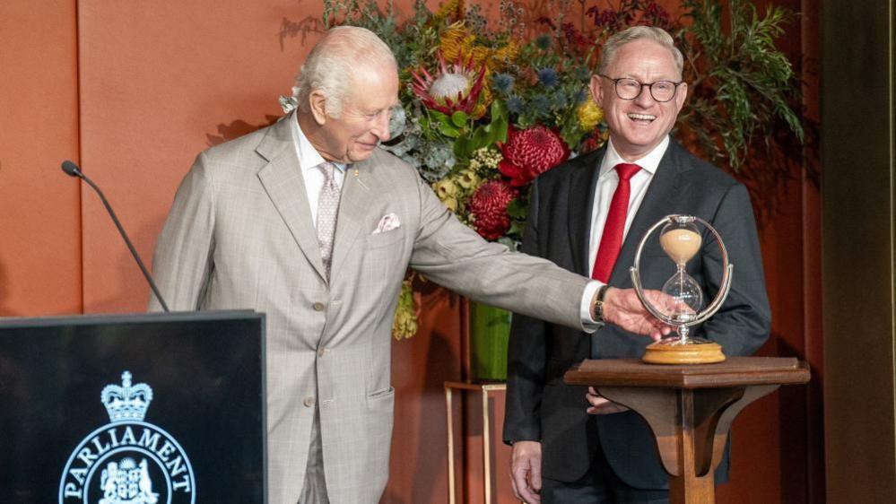 King Charles, pictured on the left, turns an hourglass that is stood on a small, high table in front of him. President of the New South Wales Legislative Council, Ben Franklin is on his right, he can be seen smiling and looking out towards the crowd.

Charles is wearing a light grey suit and Franklin is wearing a grey suit with a red tie.