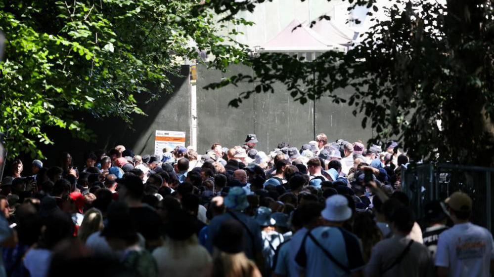 A large crowd are waiting outside the gate to the park. Many are wearing bucket hats.