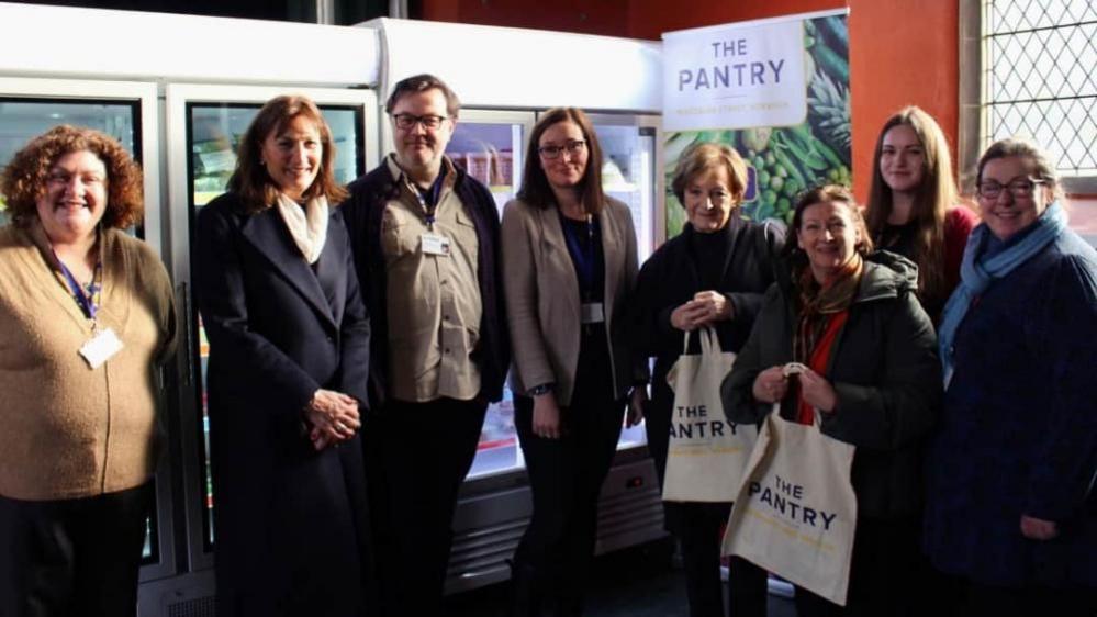 Delia Smith stands fourth from right in an image where people gathered for the opening of The Pantry in St Saviour's Church in Norwich. Behind the people are glass-fronted fridges and a banner stating "The Pantry" stands behind Delia. A leaded window is seen on the right of the image.