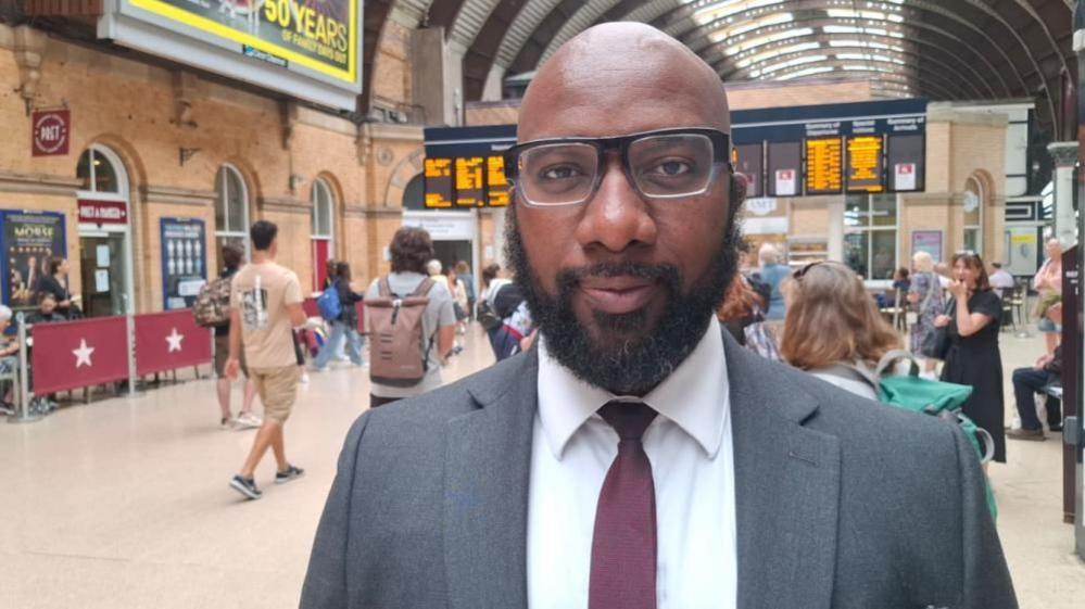 A man with a dark beard, and wearing glasses and a suit and tie, looks into the camera. He is standing in York Railway Station with the departure board behind him. People are walking around in the background. 