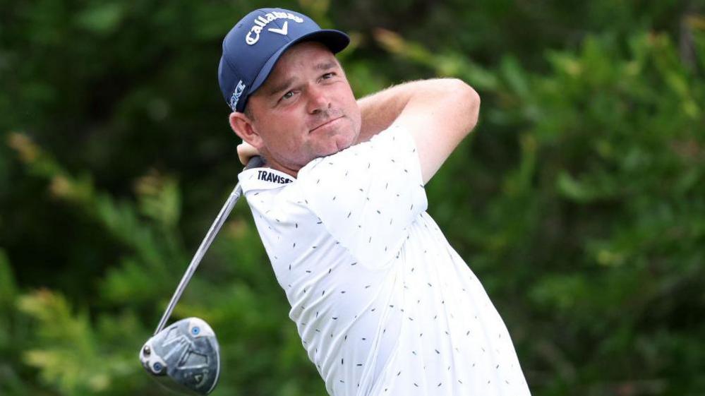 Dale Whitnell, wearing a white T-shirt, watches his ball after taking a shot with his driver