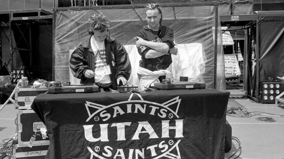 Two men stood behind decks on a stage. The deck table has a sheet over it with writing which says Saints Utah Saints.