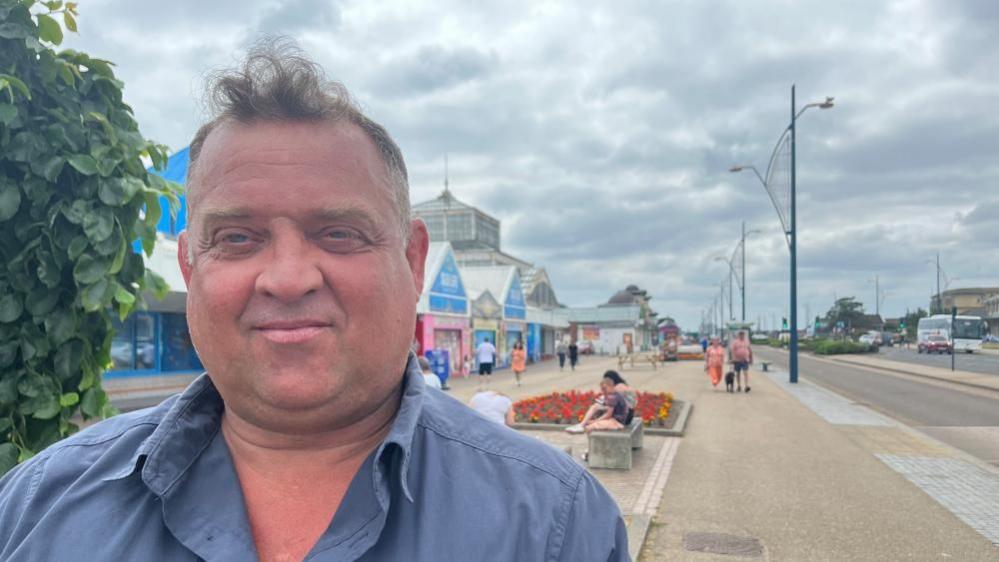 Asa Morrison, with combed receding hair, is tanned and standing on the Great Yarmouth promenade. He is wearing a blue shirt and behind him is the Great Yarmouth Sea Life Centre, and the Winter Gardens.