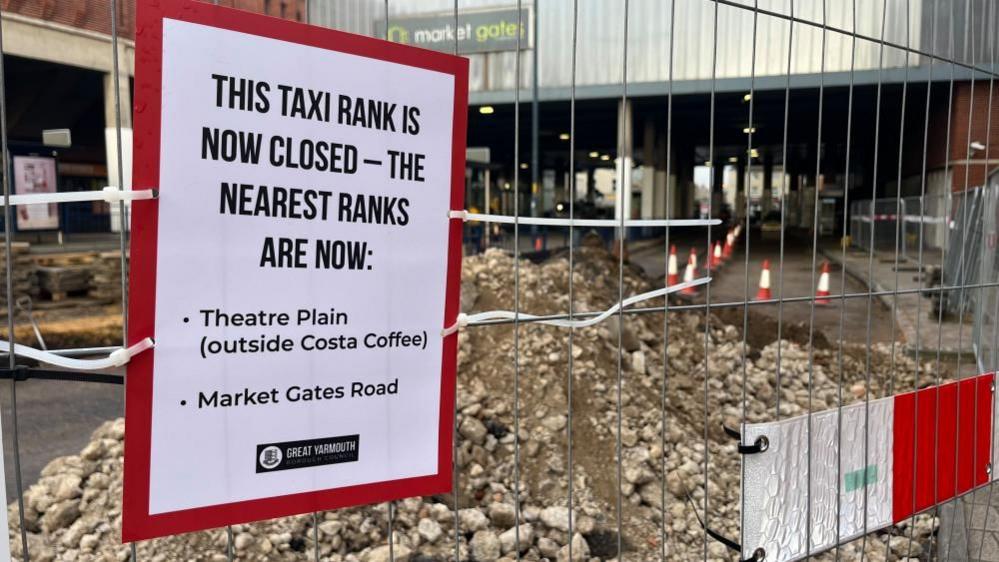 A sign pinned onto temporary fencing at Great Yarmouth Market Gates bus station on Temple Road, reads: This taxi rank is now closed - the nearest ranks are now: Theatre Plain (outside Costa Coffee) and Market Gates Road. Behind the fencing is a pile of rubble on the roadway and the Market Gates shopping centre.