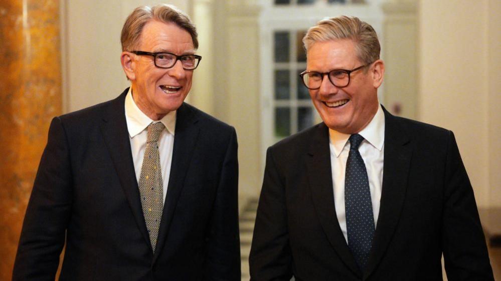 Lord Mandelson is walking alongside Sir Keir Starmer, with both men wearing dark suits, glasses, white shirts and ties with a pattern on them. There is a white framed door with glass panels behind them.