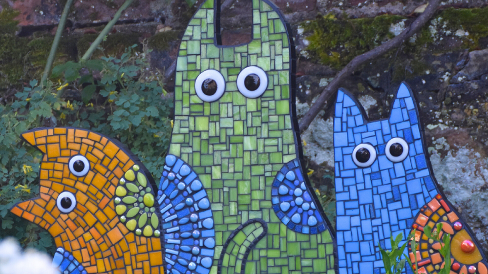 Three stylised cat-shaped mosaics in blue, green and orange with googly-type eyes. They have an old wall behind them