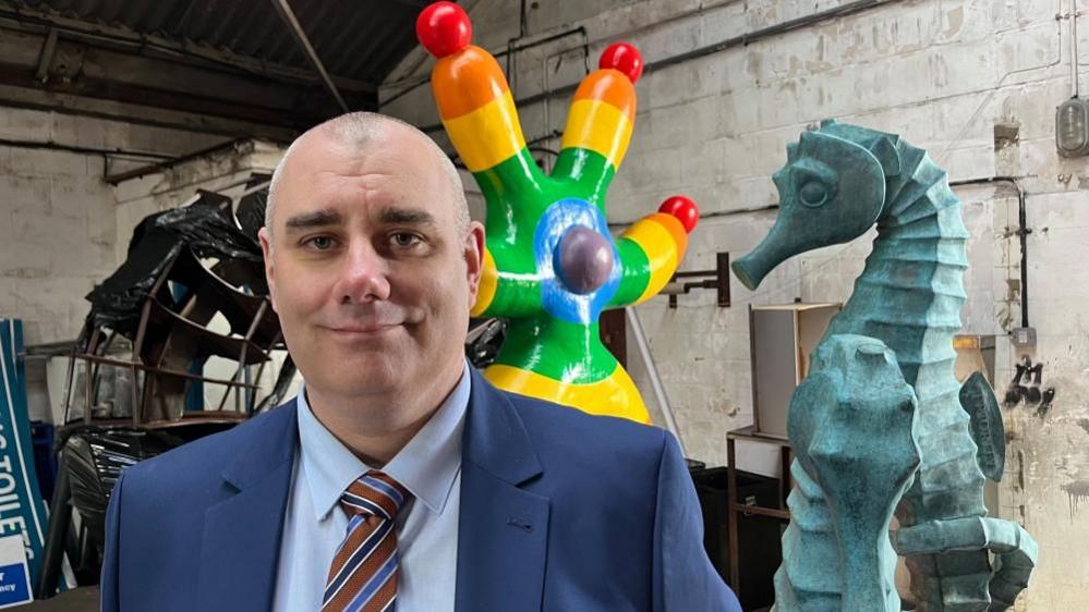 Daniel Candon has a close cropped head of hair. He is wearing a blue suit, a light blue shirt and a red tie with light and dark blue, and white, stripes diagonally across the design. He is standing in a warehouse with several sculptures around him.