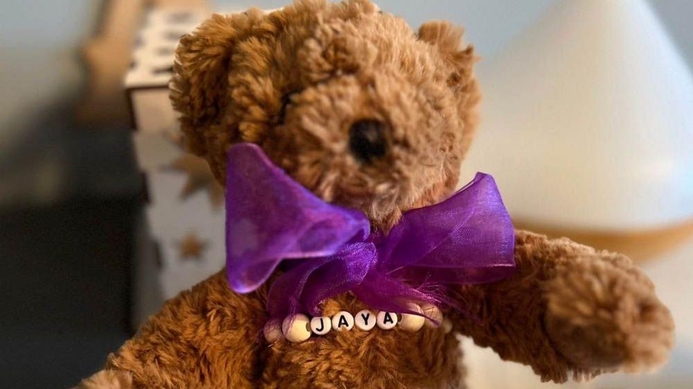 Close up of a small brown teddy bear with a purple bow round its neck and a beaded chain across the chest saying 'Jaya'