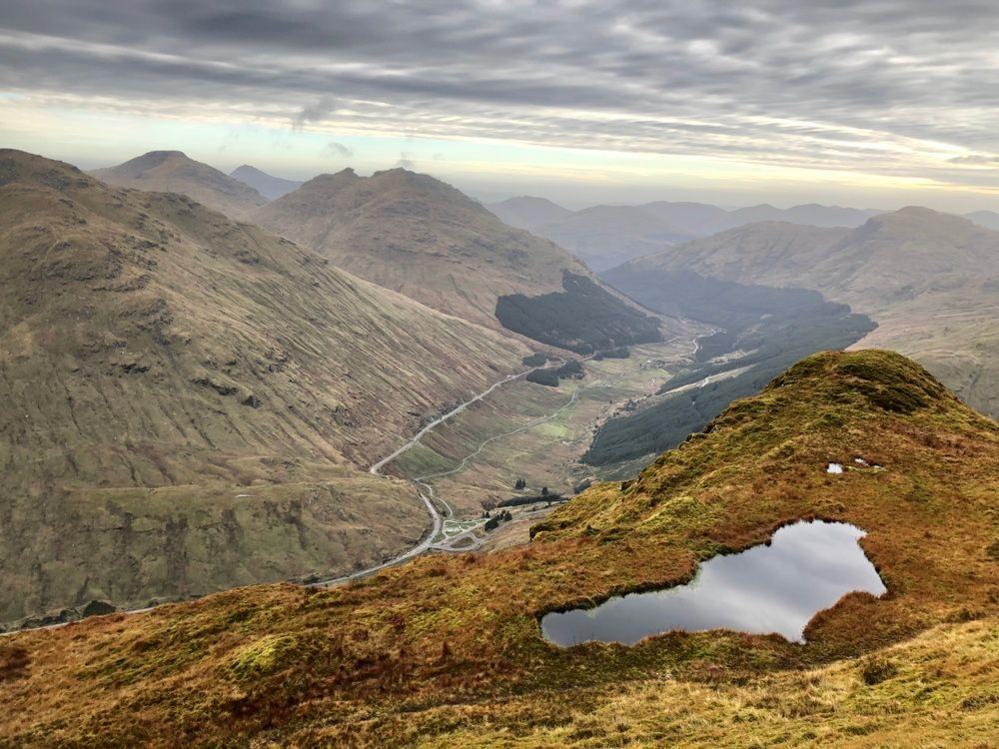 The view of the Rest and Be Thankful road winding around hillsides taken from Beinn an Lochain. The hillside is mostly brown with patches of greenery. A body of water is on top of the hill the photographer stands on.