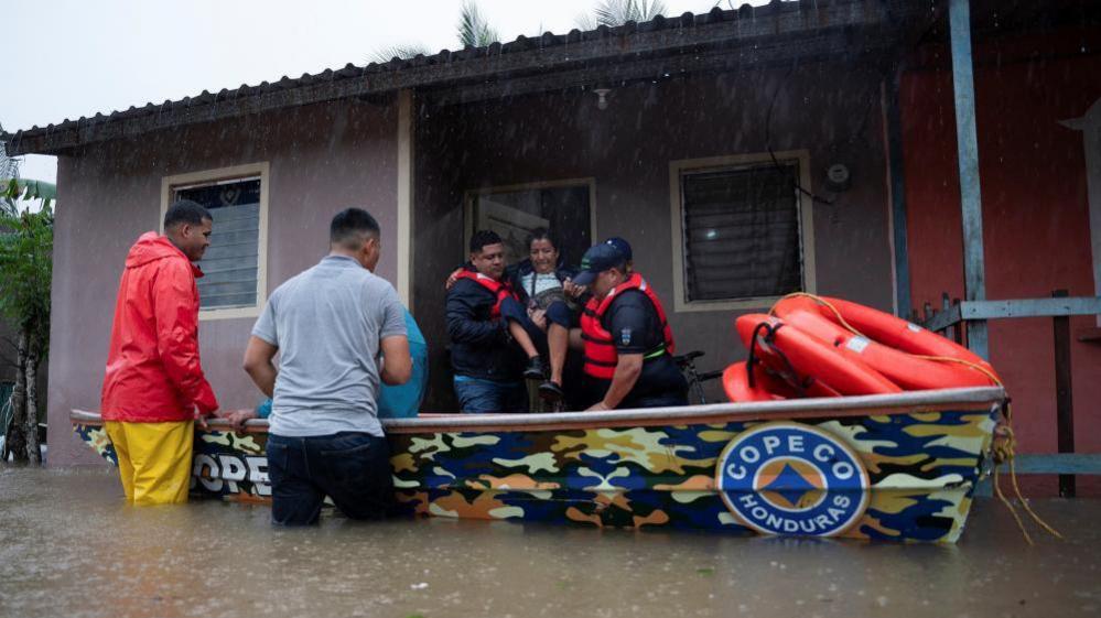 Rescue workers wearing red inflatable jackets carry a woman aloft into a waiting boat. Rain is falling and the road is flooded with water reaching the rescuers' thighs. 