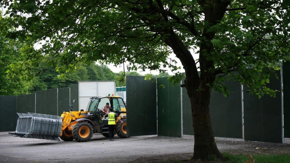 A person driving a forklift carrying metal barricades drives out between large barriers in a park.