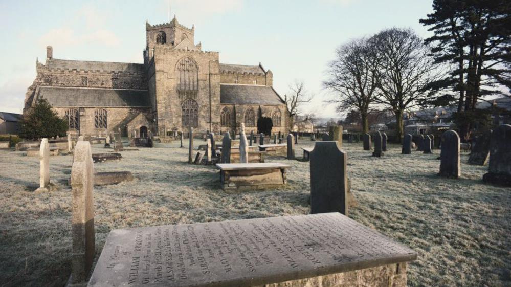 A general view of Cartmel priory church and surrounding graveyard, circa 1980. The graves are in the foreground with the priory behind. The stone, gothic construction has a square central tower set at an angle to the main building.