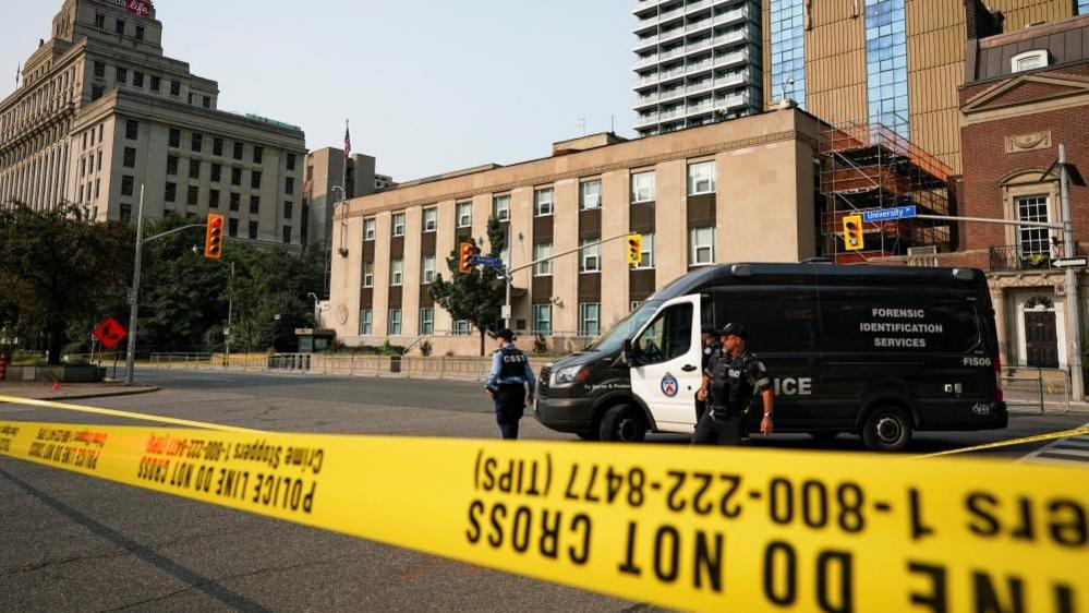 An image showing two Toronto Police officers standing behind yellow caution tape across a three-lane road. Behind them is a black and white Forensics Investigation Services van. The US consulate building, which is beige stone, is seen in the background. 