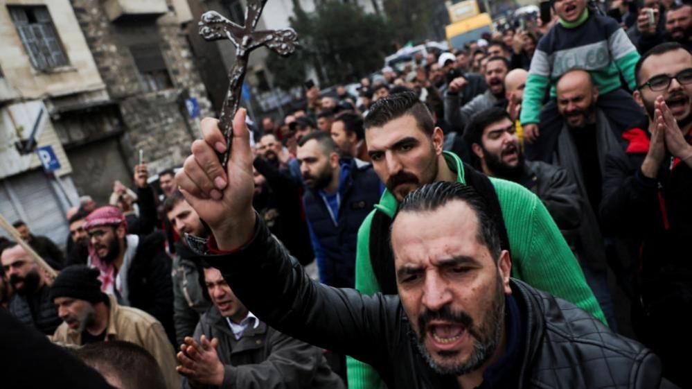 A shot of a crowd of protesters marching through a street. In the foreground, a bearded man holds up a small cross as he yells.