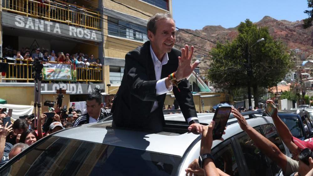 Jorge Quiroga stands up through a car's sunroof and waves to crowds.