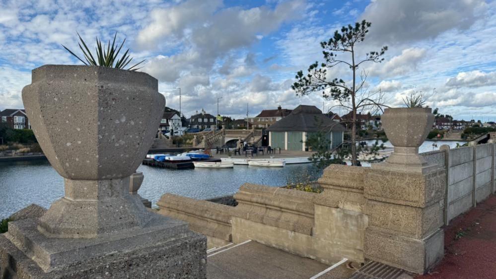 Two concrete runs with planting create the entrance to the Boating Lake, down a set of steps. Beyond is the water, where boats are stacked on the hardstanding, and the thatched cafe building is beyond.