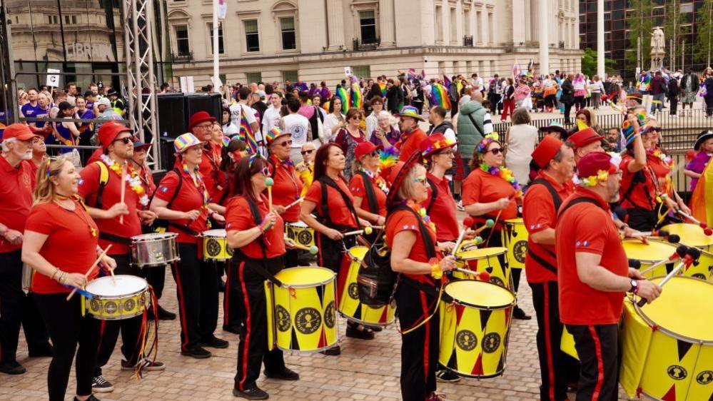 A group of drummers who are part of a Pride Parade. They are in the middle of a city centre and are wearing black trousers with a red stripe and red t-shirts. 