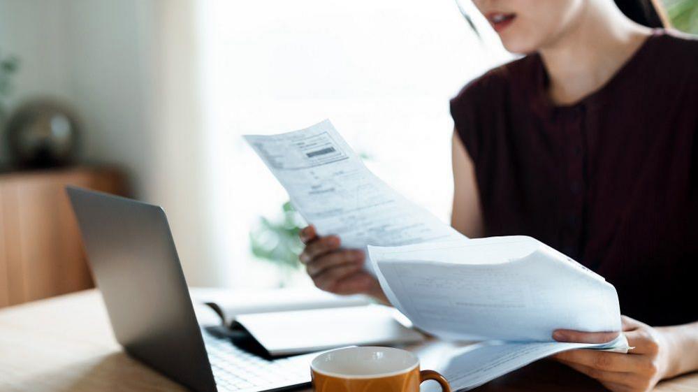 Woman blurred at coffee table looking at bills in front of a laptop. 