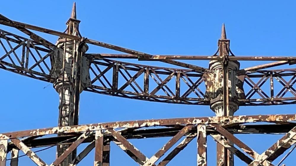 The Great Yarmouth gasometer stands in a blue sky, with it's peeling paint and rusting of the columns and lattice work. With finials on the top it is is highly decorated, with it being awarded Grade II listed status.