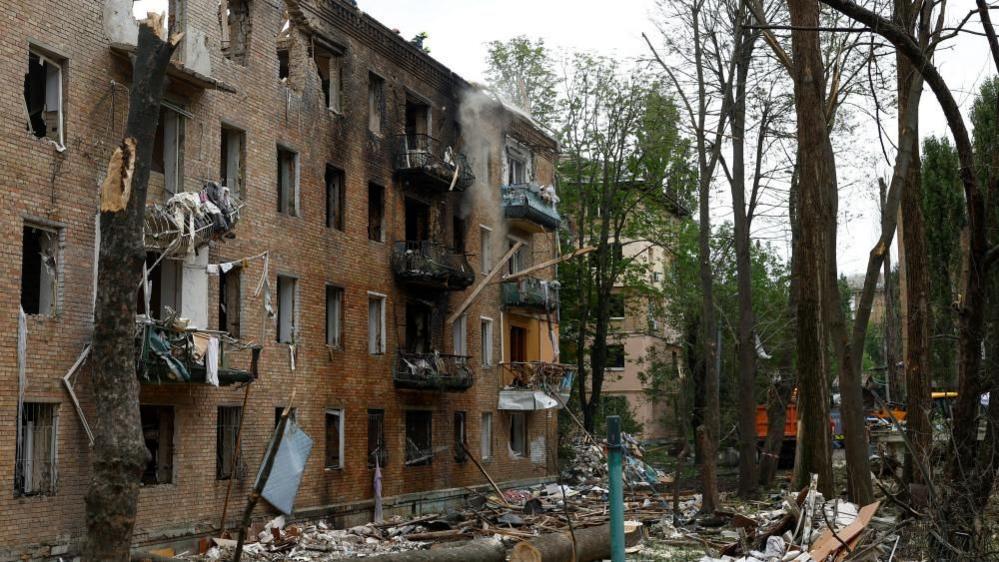 The image shows a red bricked building obliterated from a missile and drone attack, balcony's and windows blasted out and shrapnel and debris all over the floor surrounded by trees