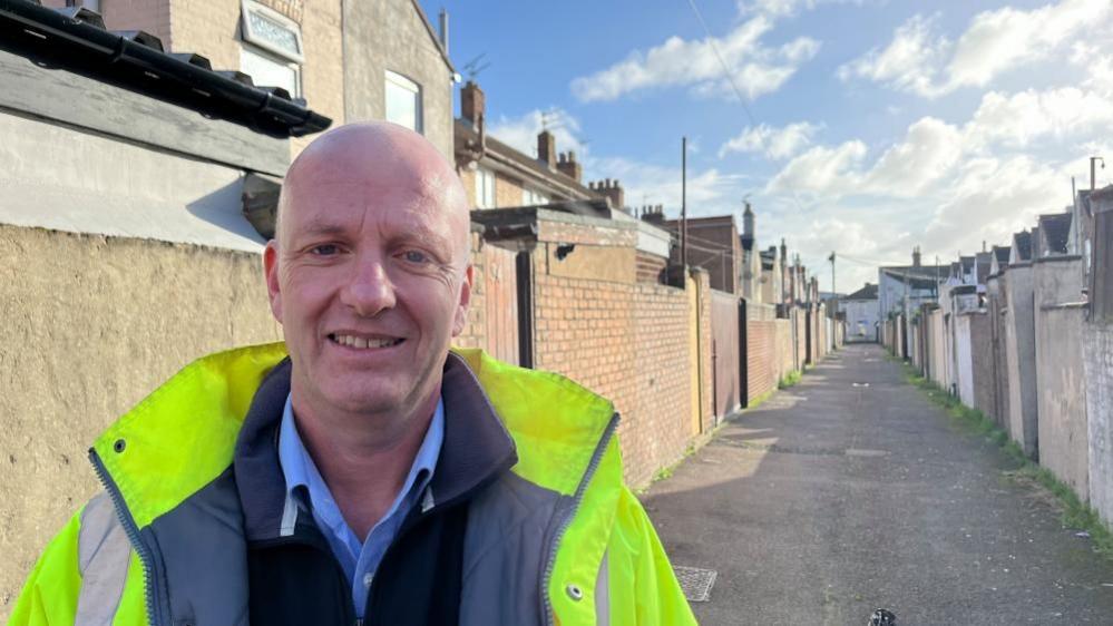 James Wilson has a clean shaved face and head. He is standing in an alleyway, with the backs of properties visible behind him, and he is wearing a high visibility yellow jacket, with silver reflective stripes, and a fleece and blue shirt underneath.