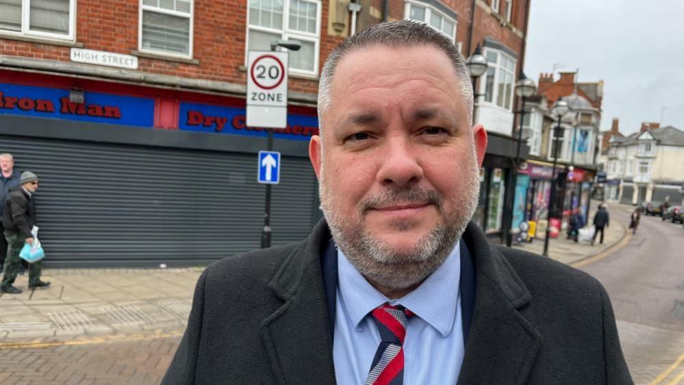 Jason Smithers looks directly at the camera as he stands in a street, with short dark hair and beard and wearing a shirt and tie. 