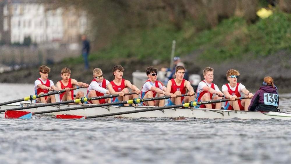 8 young men are pictured in a rowing boat, mid-race. All are wearing red shorts and vests, with some wearing a white top underneath and others wearing sunglasses. A Cox with a purple hoodie is pictured at the rear of the boat. They are all reaching forward with their oars in preparation for a stroke.