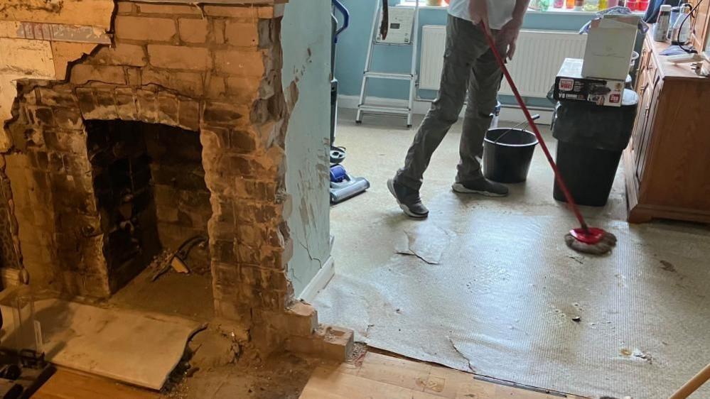 A view inside Mrs Richardson's home while her husband mops the kitchen floor, with the damaged fireplace in the foreground.