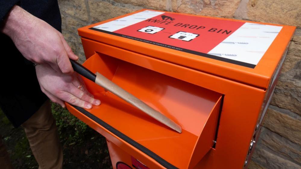 A person's hand can be seen opening the slot of an orange bin and putting in a knife. The blade of the knife is covered in carboard. 