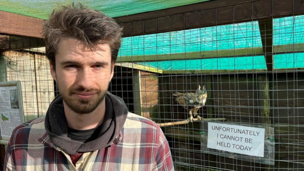 Adam Brunger is wearing a lumberjack shirt, with a black hoodie and black teeshirt underneath. He has a full but short beard and moustaches, and has long dark hair. He is standing next to an owl enclosure, with an owl in the background, and a sign reading: "unfortunately I cannot be held today".